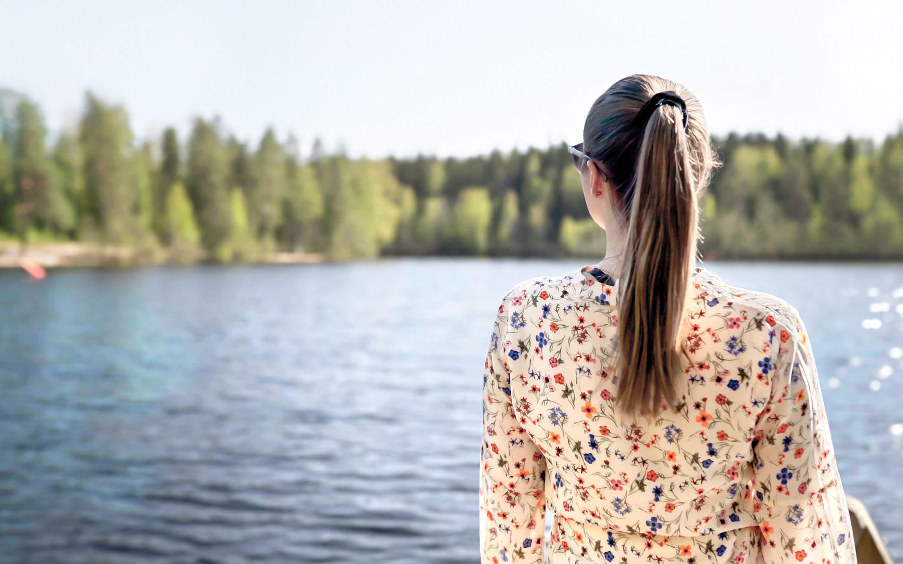 Eine Frau in einer Bluse mit Blumenmuster schaut auf einen See. Das Ufer grenzt an einen Wald voller Bäume.